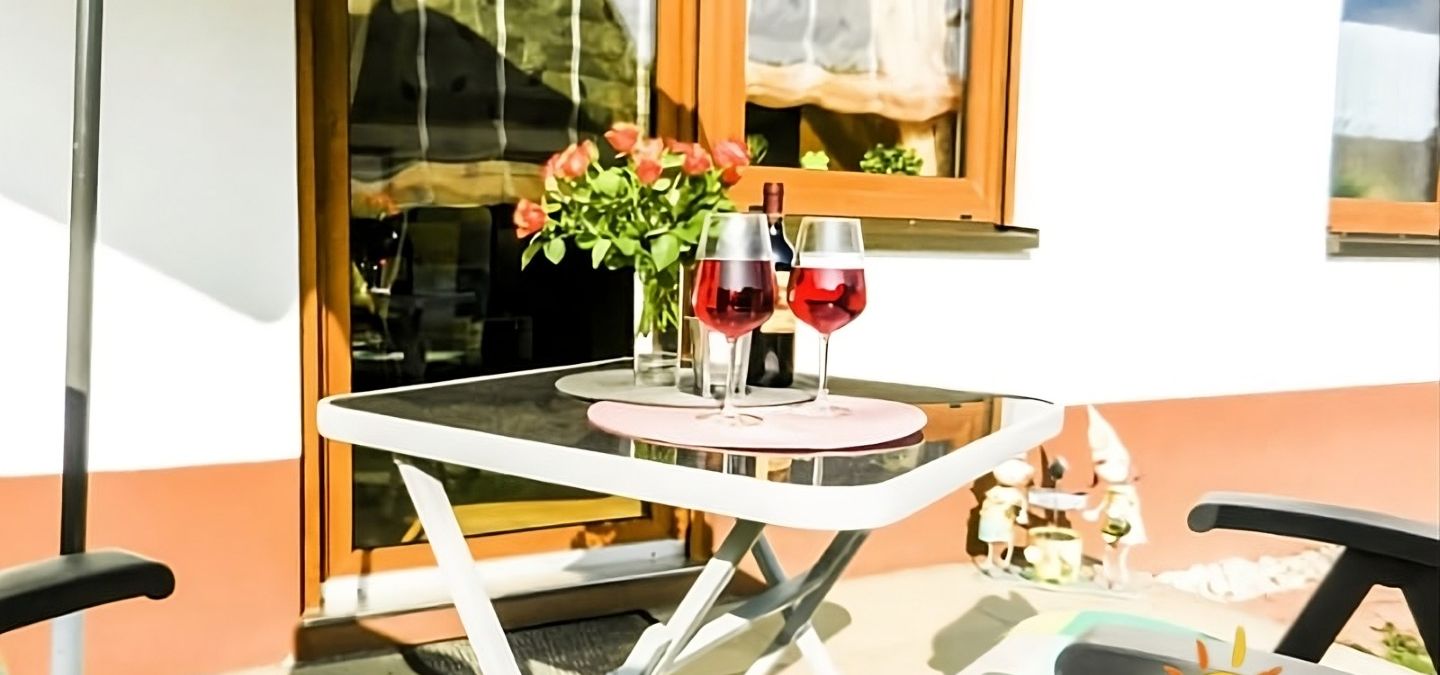 Gemütliche Terrasse der Ferienwohnung Ilona in Zweibrücken mit zwei Gläsern Roséwein, Blumenarrangement und bequemen Gartenstühlen – perfekter Ort zum Entspannen am Abend.